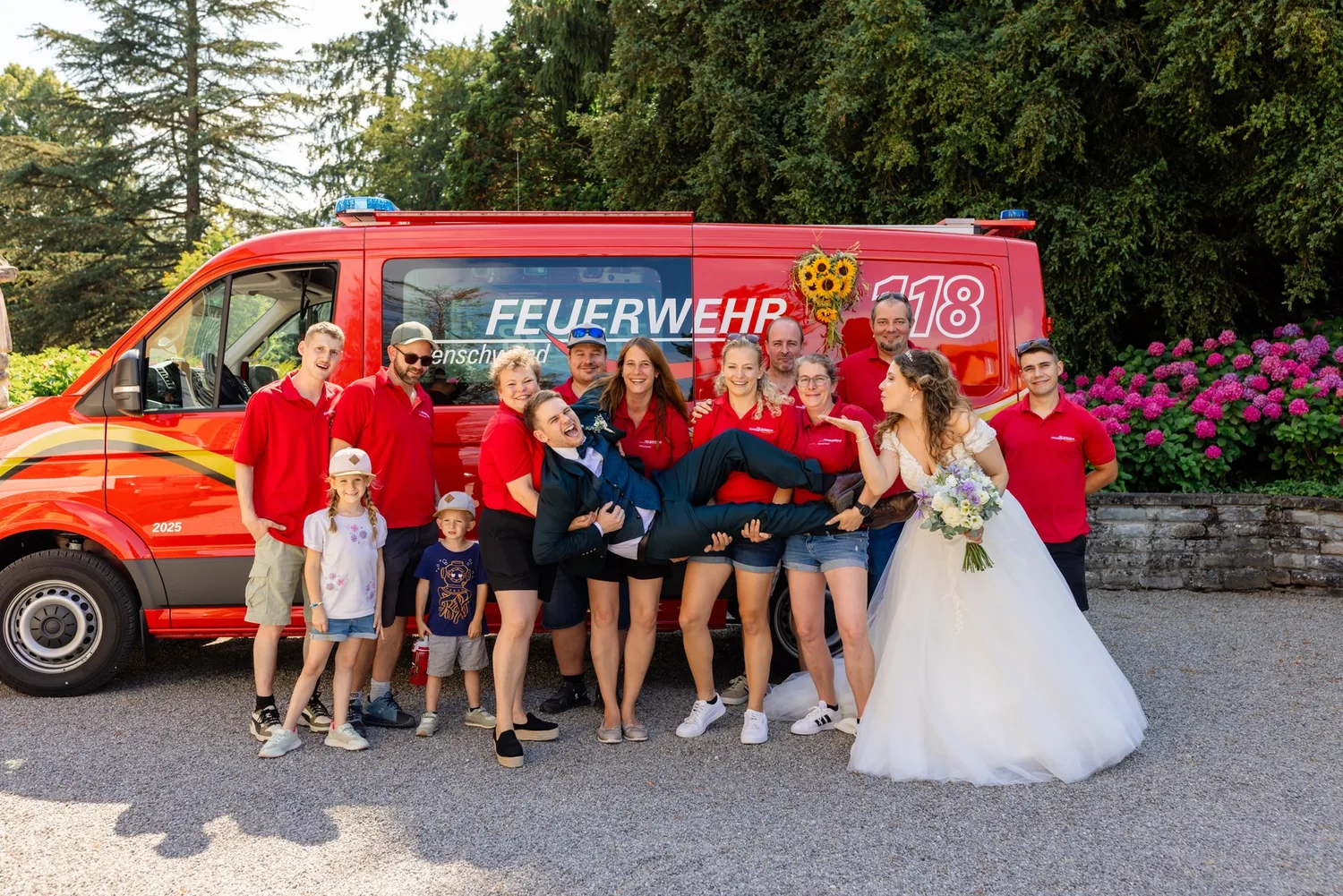 Feuerwehrleute in roten Shirts vor ihrem Fahrzeug tragen den Bräutigam, daneben die Braut