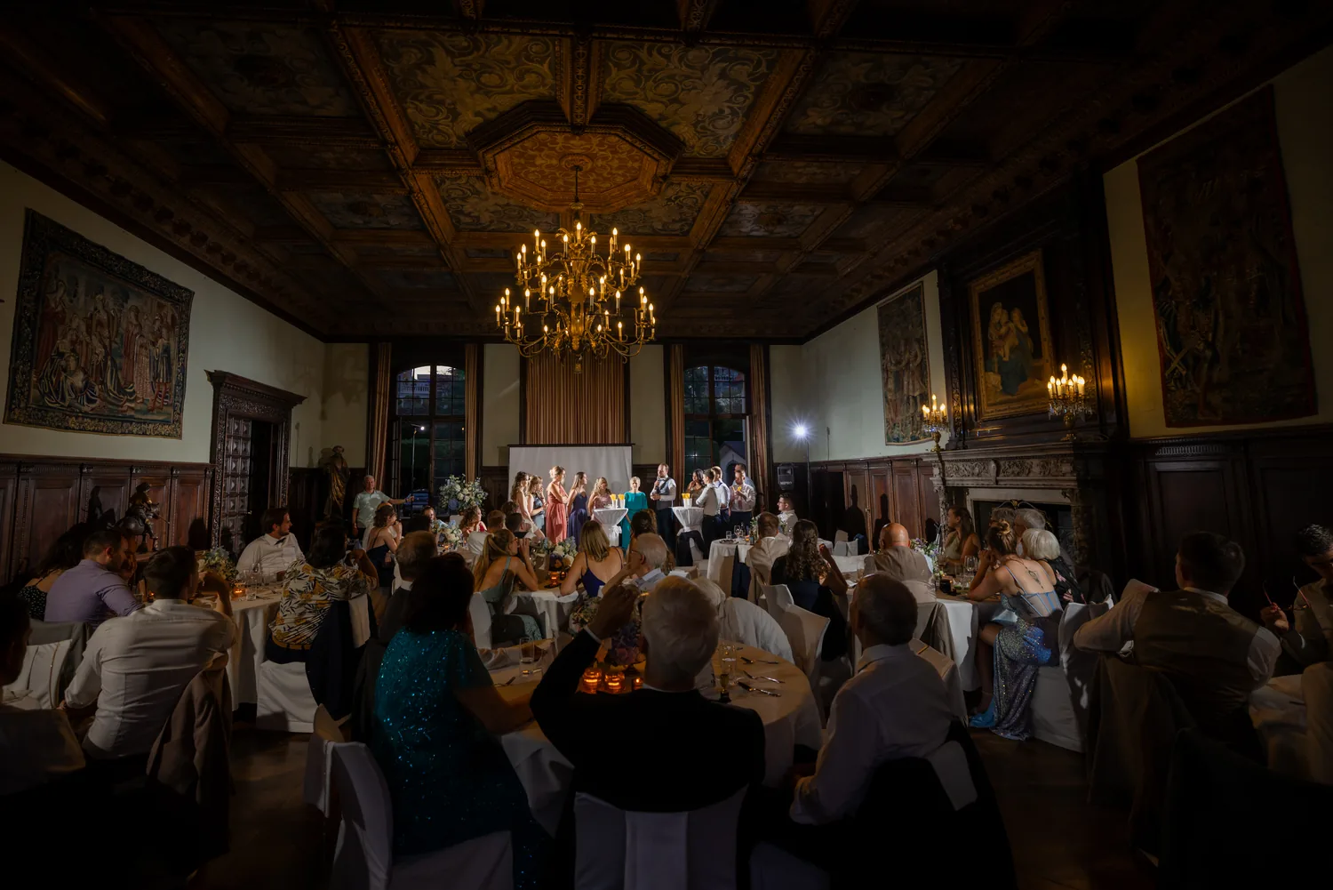 Blick in den Rittersaal beim Dinner: bemalte Kassettendecke, Kronleuchter, Gobelins an den Wänden, die Gäste an runden Tischen, vorne eine Darbietung