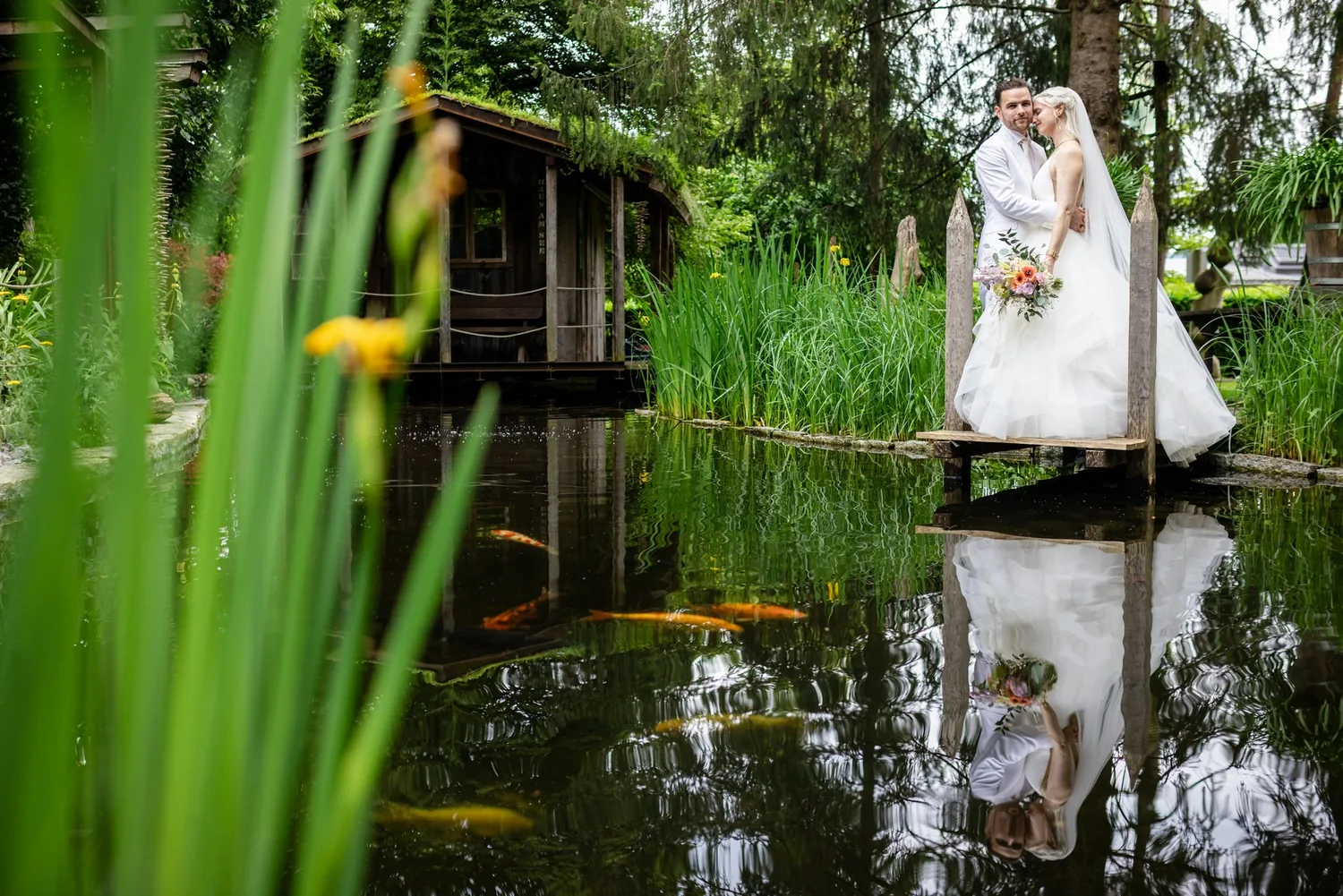 Brautpaar steht auf einem schmalen Holzsteg über dem Teich, im Wasser spiegeln sich beide