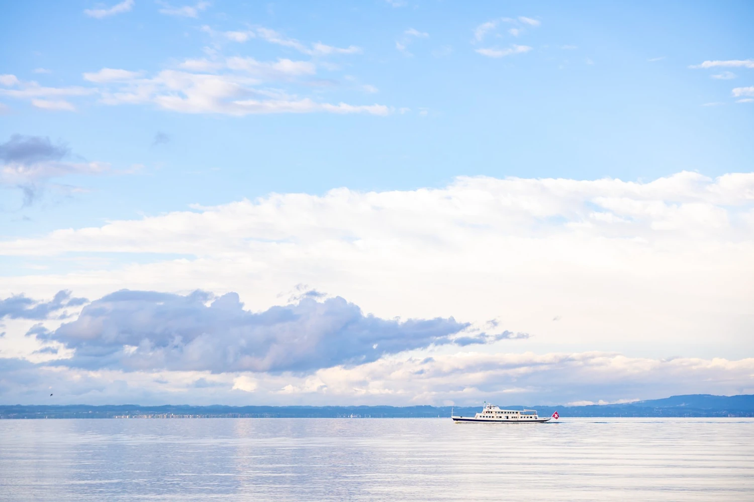Blick über den Bodensee mit einem Kursschiff und dem gegenüberliegenden Ufer unter Wolken