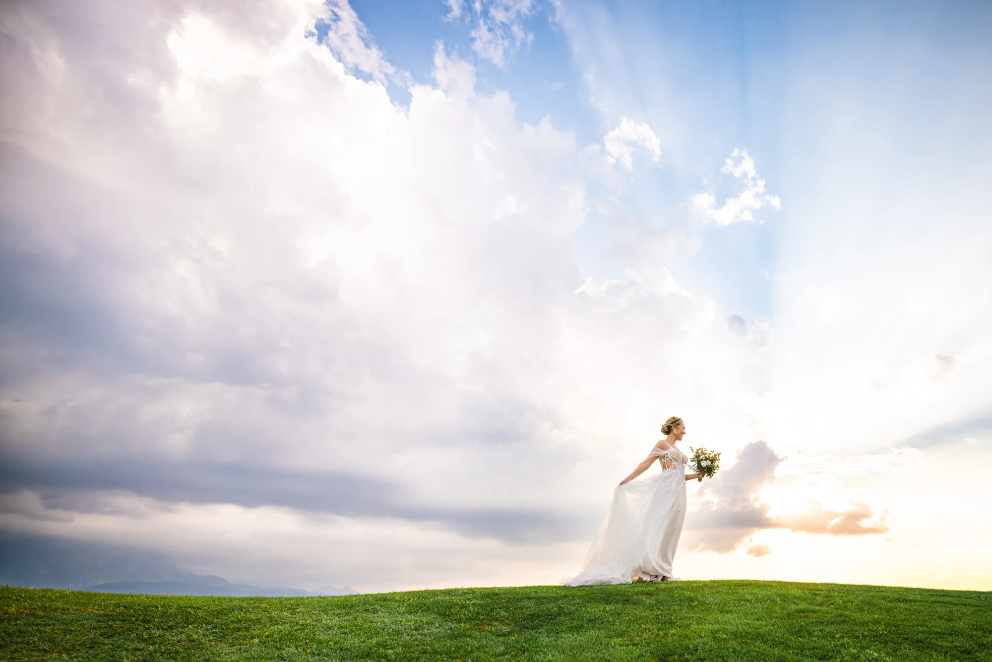 Braut im langen Kleid steht mit Brautstrauss auf einer grünen Hügelkuppe, über ihr ein weiter Wolkenhimmel mit Sonnenstrahlen