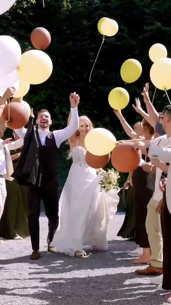 Brautpaar geht Hand in Hand durch ein Spalier aus Gästen, die gelbe, weisse und terrakottafarbene Luftballons in die Höhe halten