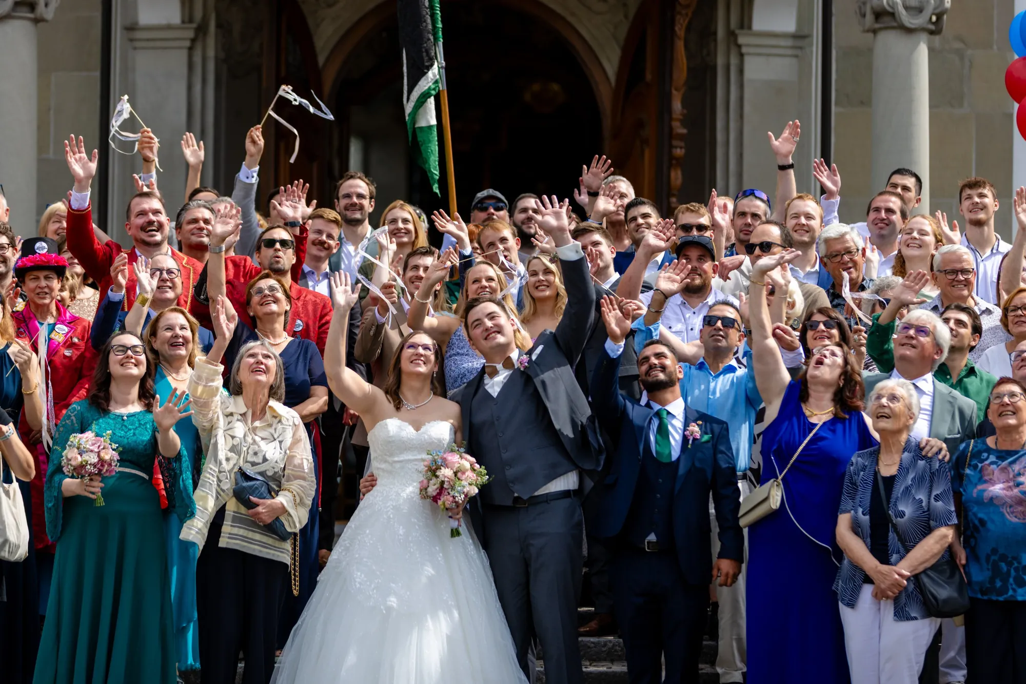 Brautpaar und Hochzeitsgäste winken mit Bändern in die Luft, vor dem Portal einer Kirche