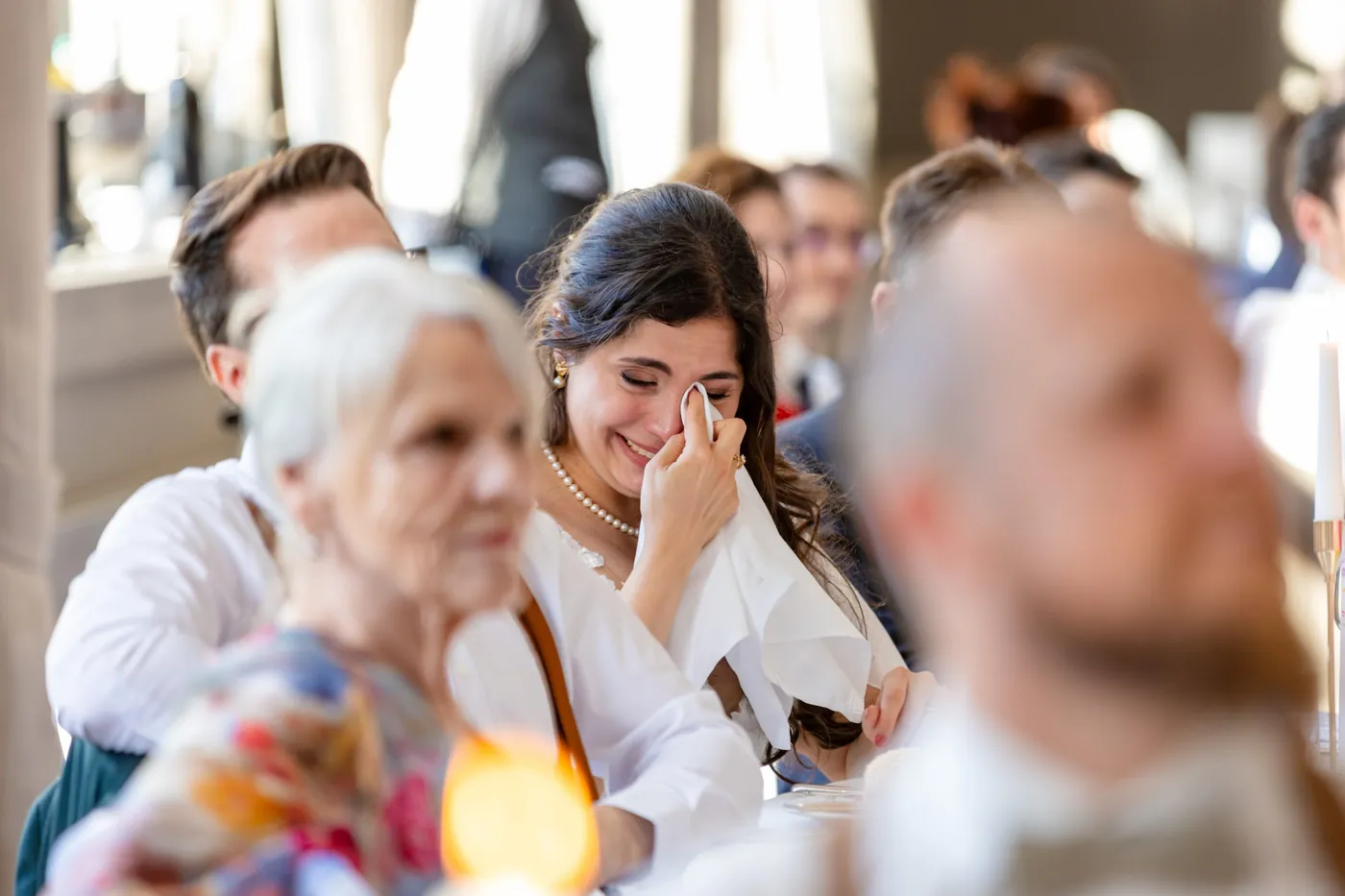 Die Braut sitzt am Tisch und wischt sich während einer Rede lachend die Tränen ab