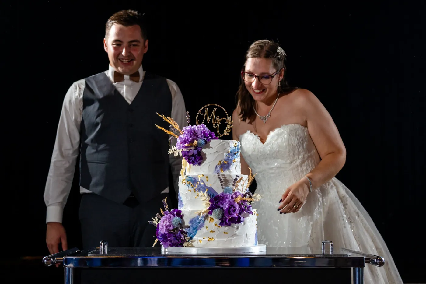 Das Brautpaar steht lachend vor der dreistöckigen Hochzeitstorte mit violetten Blumen